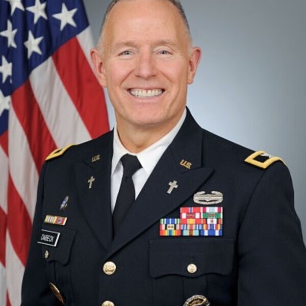 Lawrence Dabeck headshot. A man in U.S. Army dress uniform stands smiling before an American flag. He wears a black tie and numerous decorations and medals are pinned to his jacket. Two cross pins indicate his role as a chaplain.