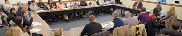 NDISC welcomes prominent speakers to Notre Dame to discuss International Security topics. A group of people sit around a square-shaped ring of tables in a meeting room, engaging in a discussion. Laptops and notebooks are open on the table, suggesting an active work session.