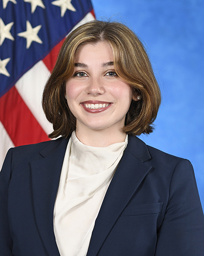 Smiling woman with shoulder-length brown hair, wearing a navy blue blazer and cream blouse. US flag in background.