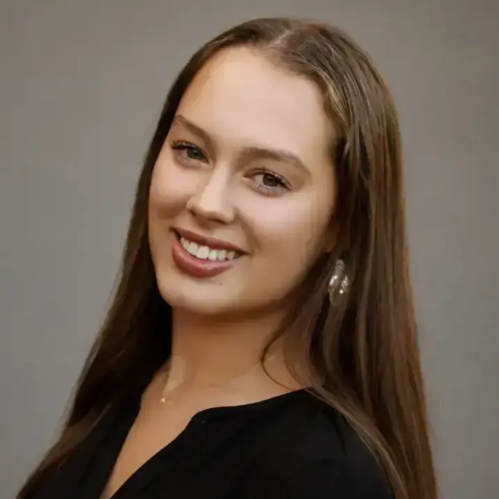Smiling young woman with long brown hair and green eyes, wearing a black top, a gold necklace, and large pearlescent earrings.